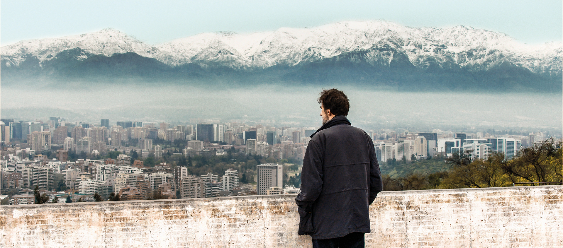 a man in a black coat overlooking a city with mountain in the background