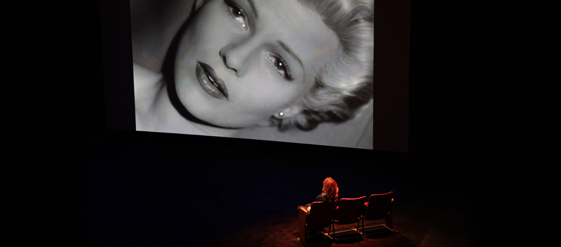 Nina Menkes sits on stage and watches a black and white film scene that depicts a woman's face up close.