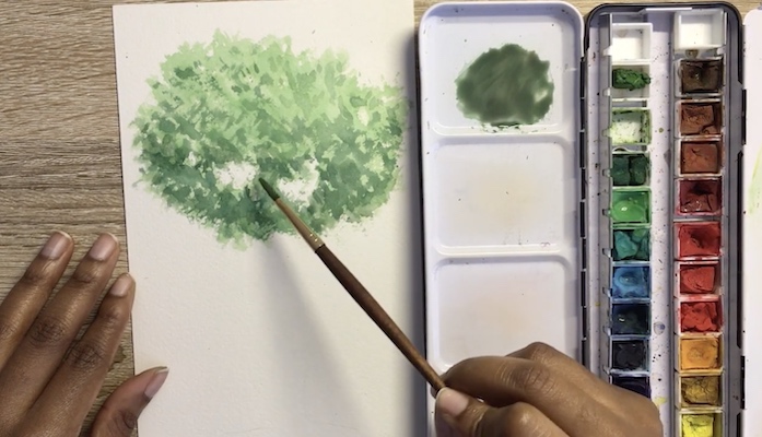 On a wood work surface with an open container of watercolor paints, a black woman's hands apply green paint to white paper, creating the look of a tree top