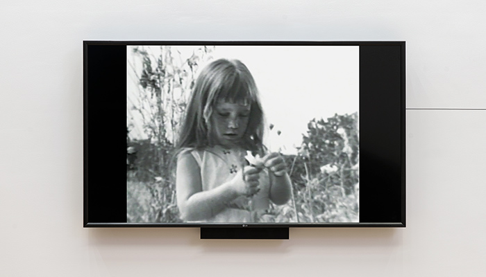A tv mounted to a gallery wall displays a black and white image of a young girl holding a flower.