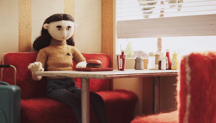 A woman character, made of felt, stares at the burger she ordered with her suitcase by her side.