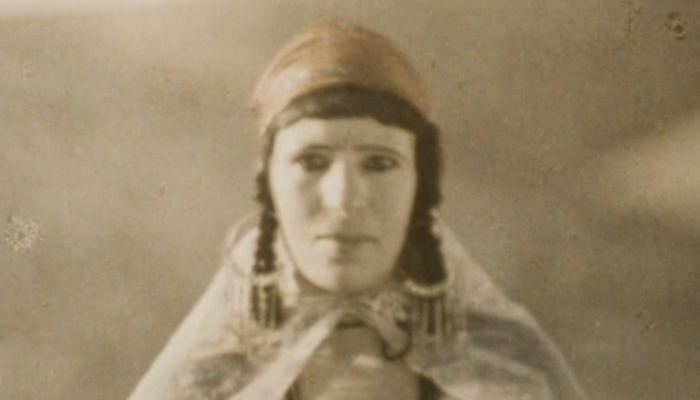 Sepia tone mage of a woman facing the camera wearing a head scarf, braids, large hoop earrings and a cape from Assia Djebar's 1982 French documentary Zerda and the Songs of Forgetting